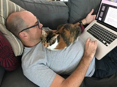 Derek Baker working on a laptop with a cat asleep on his chest